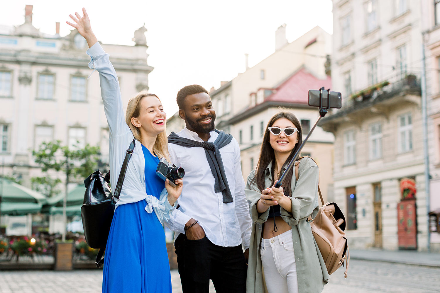 Excited cheerful mix raced friends enjoying walking outdoors in European city. Man and two women walking outside, hugging each other, laughing and making selfie photo. Multicultural friendship concept download